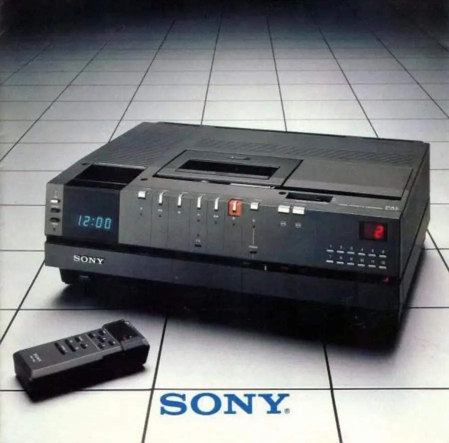 A vintage Sony Betamax VCR with a digital clock display shows "12:00" and a matching remote control in front, all placed on a tiled, grid-like floor with the blue "SONY" logo below.