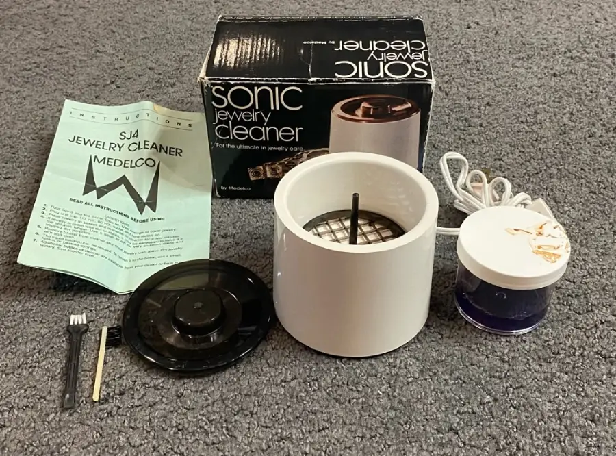 A sonic jewelry cleaner set on gray carpet, including the main device, a black lid, a brush, a manual, a small container with blue liquid, and the original product box.