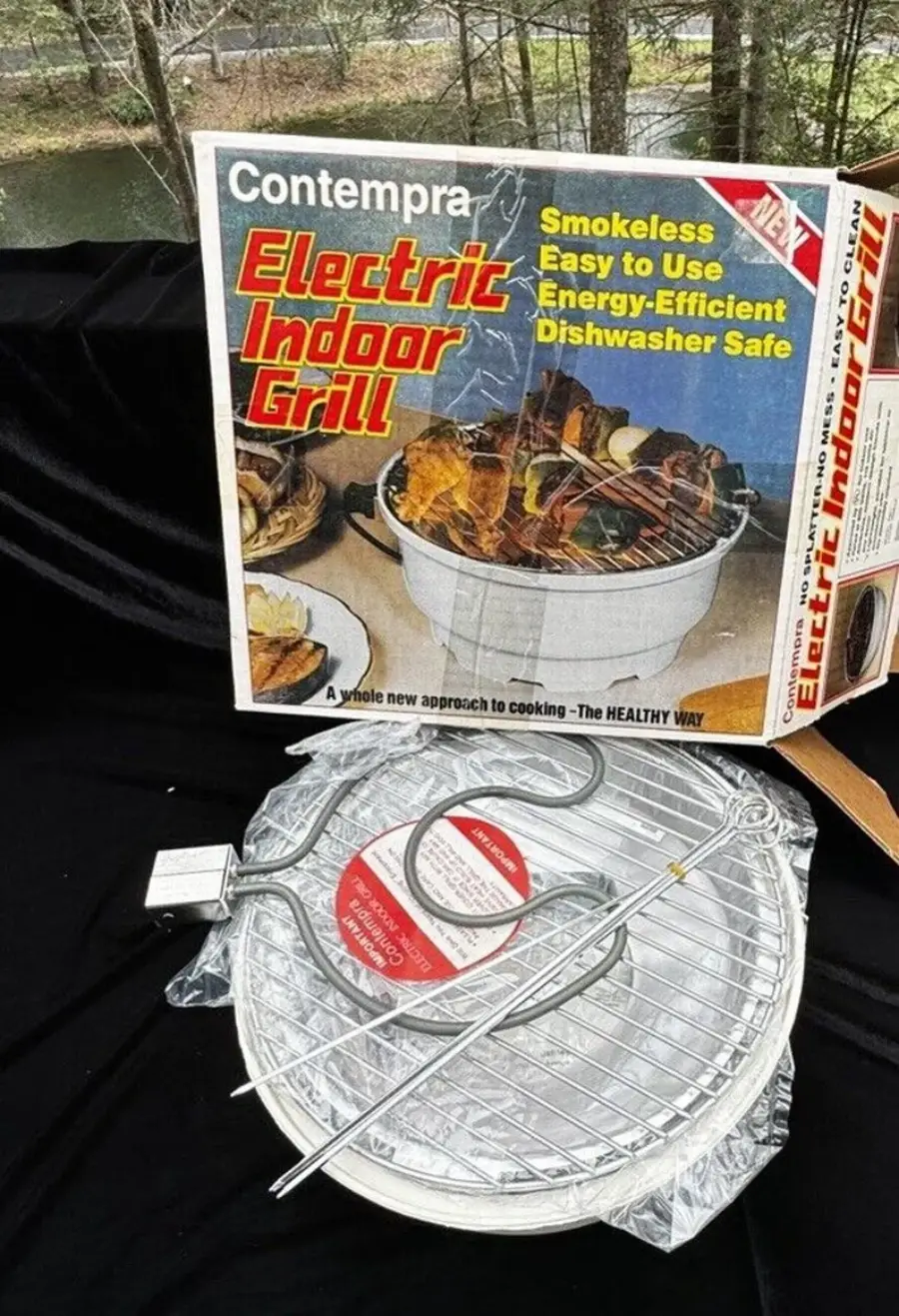 A Contempra Electric Indoor Grill set sits in front of its box, which advertises features like smokeless, easy to use, energy-efficient, and dishwasher safe. The grill components are unassembled and wrapped in plastic.