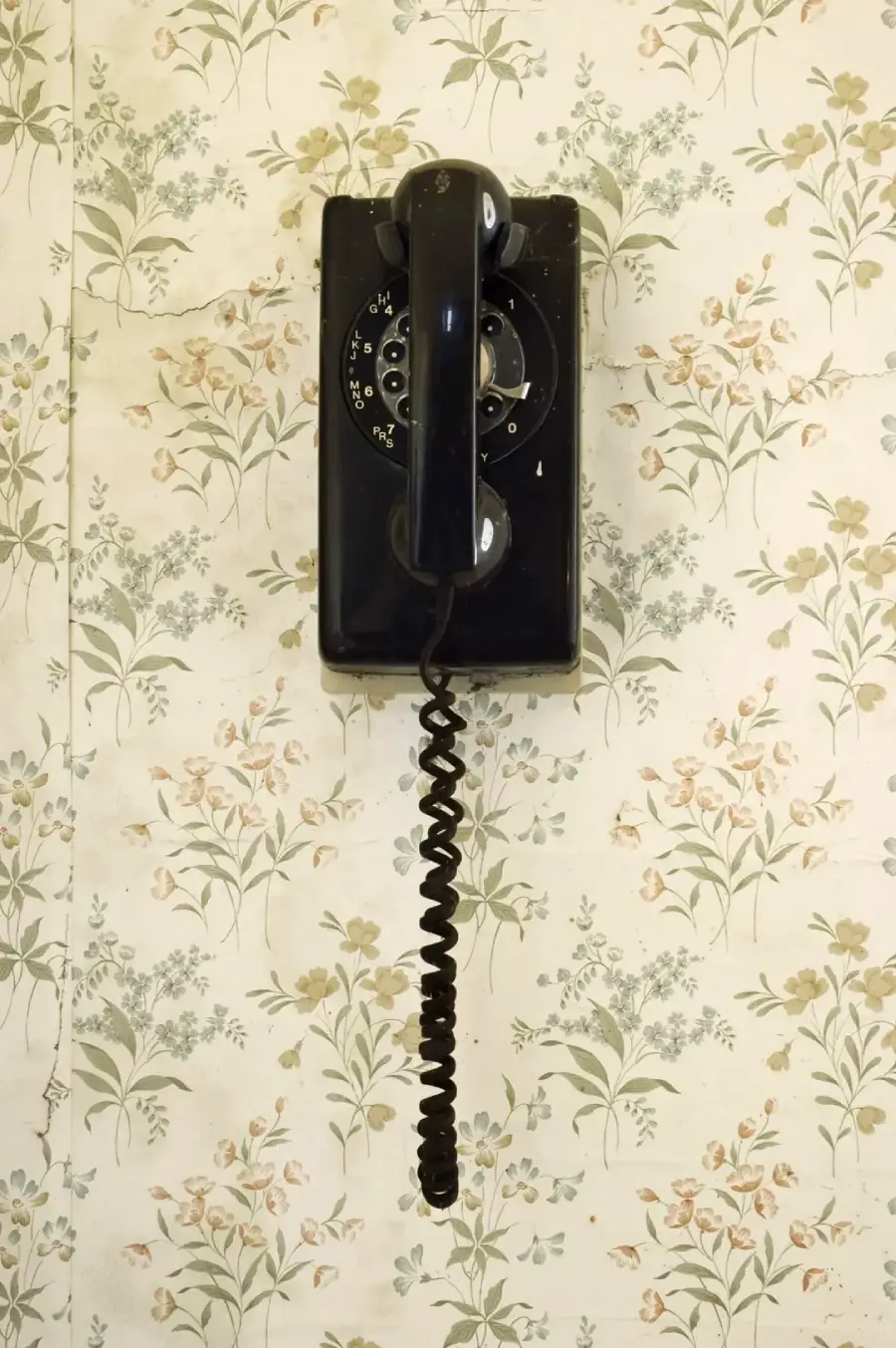 A vintage black rotary wall phone with a coiled cord hangs on floral-patterned wallpaper.