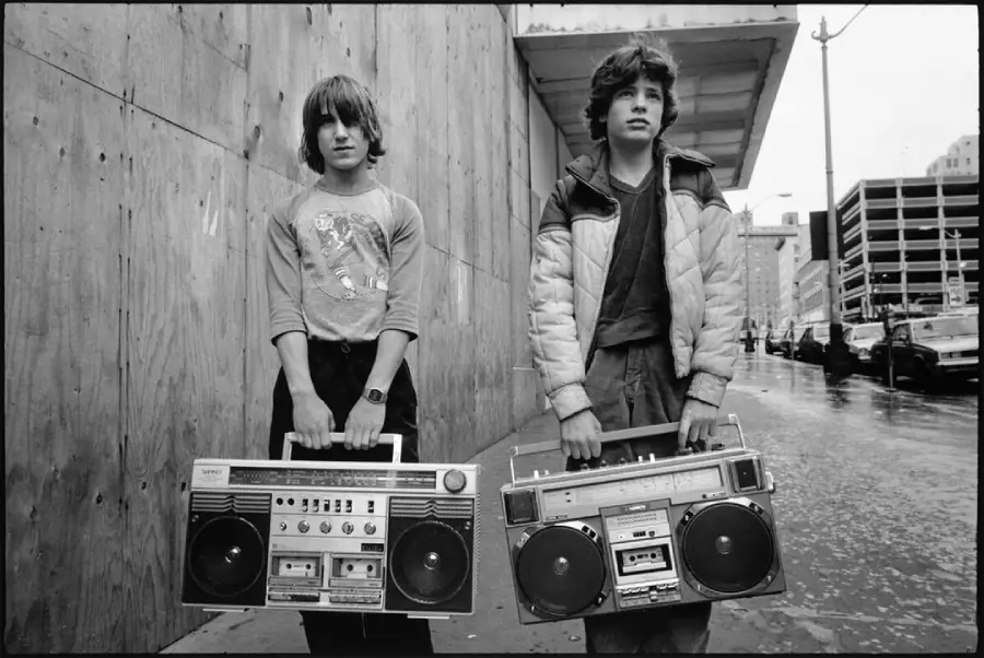 Two teenage boys stand on a city sidewalk holding large boomboxes. One wears a long-sleeve shirt and the other a puffy jacket. The background shows a wooden wall, parked cars, and wet pavement, suggesting recent rain.