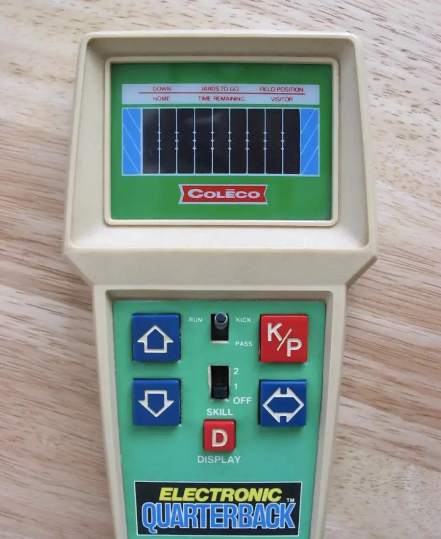 A vintage Coleco Electronic Quarterback handheld game features a green and beige plastic case, buttons for controls, and a small screen displaying a football field graphic. The device sits on a wooden surface.