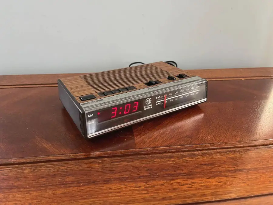 A vintage digital alarm clock with a woodgrain top sits on a wooden surface, displaying the time 3:03 in red LED numbers. The clock also has an AM/FM radio with a tuning display and control buttons.