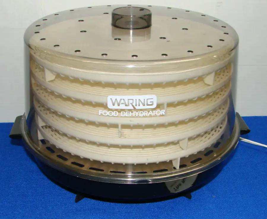 A Waring food dehydrator with a transparent plastic lid and several stacked trays is shown on a blue surface, plugged in and ready for use.