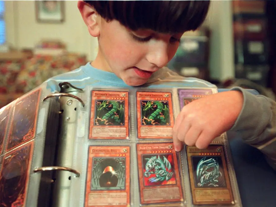 A young boy looks at and points to trading cards in a binder, showing Yu-Gi-Oh! cards displayed in clear plastic sleeves.