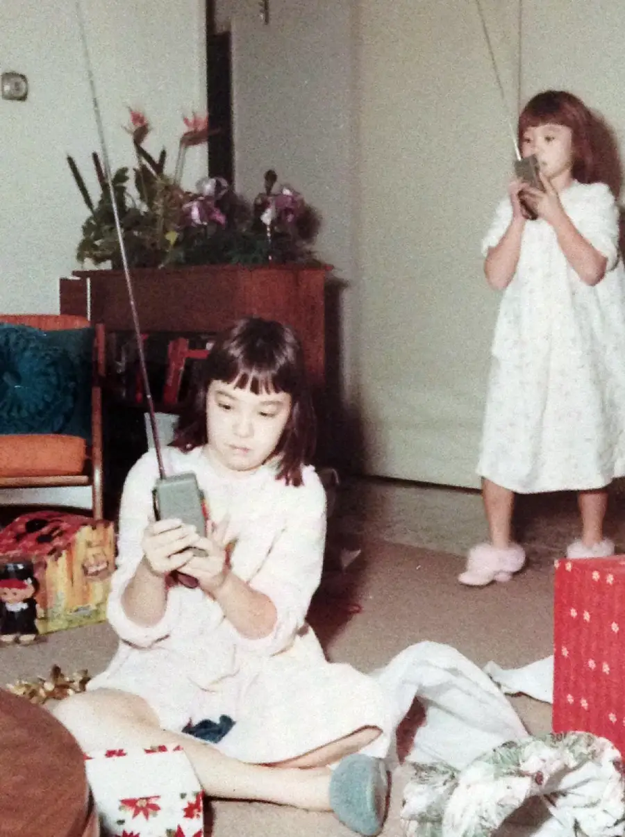 Two young girls in pajamas use walkie-talkies in a living room with gifts, wrapping paper, and toys scattered on the floor. One girl sits cross-legged while the other stands in slippers near a potted plant and a red gift box.