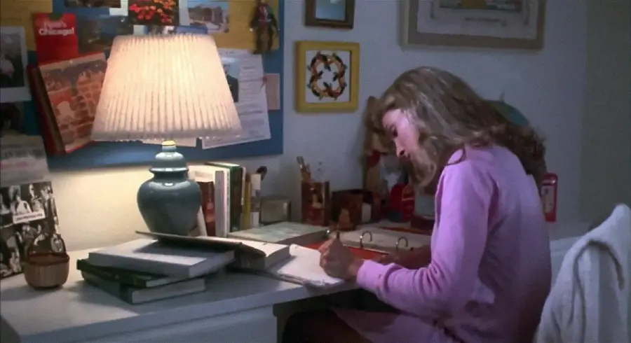 A person with long hair wearing pink pajamas sits at a desk, writing in a binder. The desk has an open book, lamp, and stacked books. The wall behind is decorated with photos, notes, and a bulletin board.