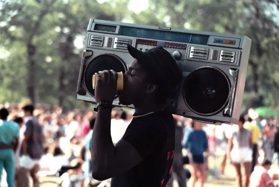 A man wearing a hat drinks from a cup while carrying a large boombox on his shoulder at an outdoor event with a crowd of people in the background.
