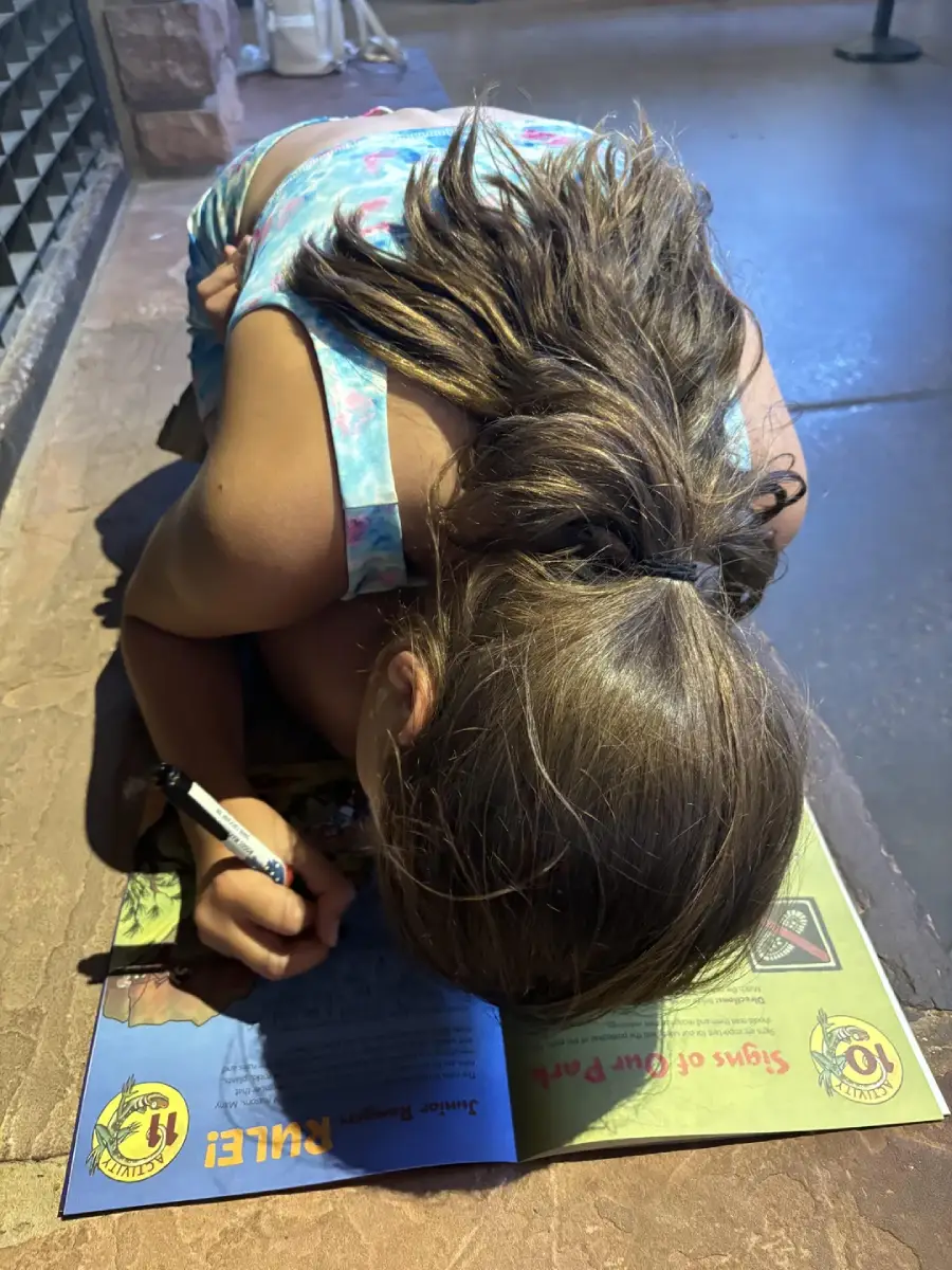 A young girl with brown hair leans over a magazine, writing with a pen while kneeling on a stone floor. The magazine features bright colors and yellow text. She wears a blue and pink tie-dye tank top.