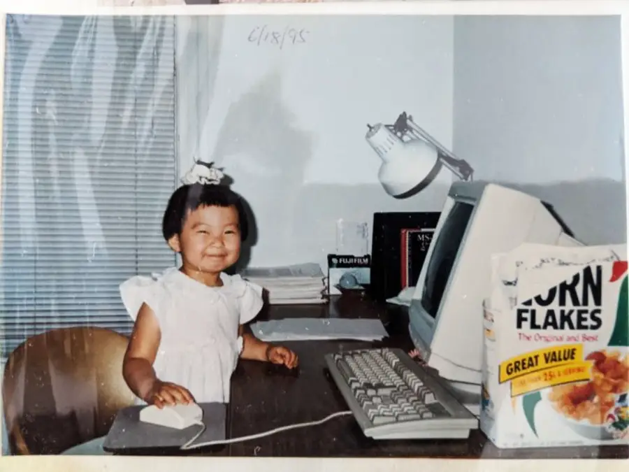 A young girl smiles at a desk with her hand on a computer mouse. There’s an old-style computer, a desk lamp, books, and a box of corn flakes beside her. The photo appears to be from the 1990s.