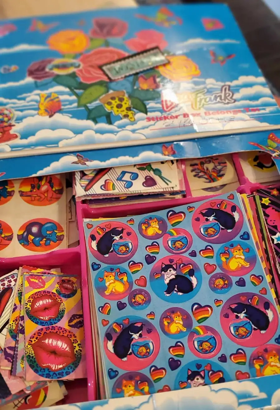 A colorful Lisa Frank sticker box is open, revealing sheets of vibrant stickers inside. The stickers feature playful cats, lips, hearts, rainbows, and other whimsical designs in bright, bold colors.