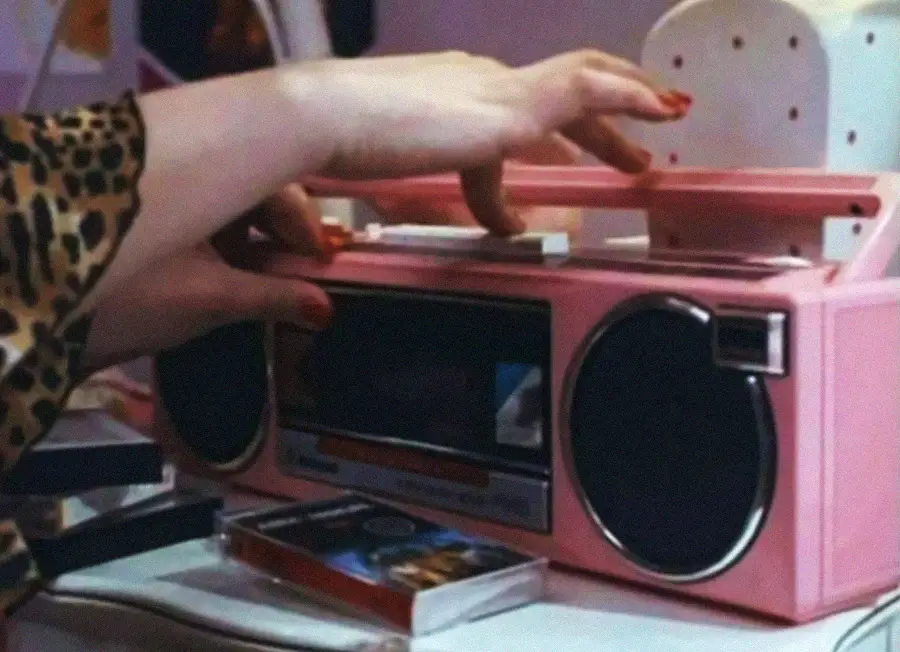 A person with red-painted nails and a leopard print sleeve is placing a cassette tape into a pink retro boombox on a desk, with another cassette lying nearby.