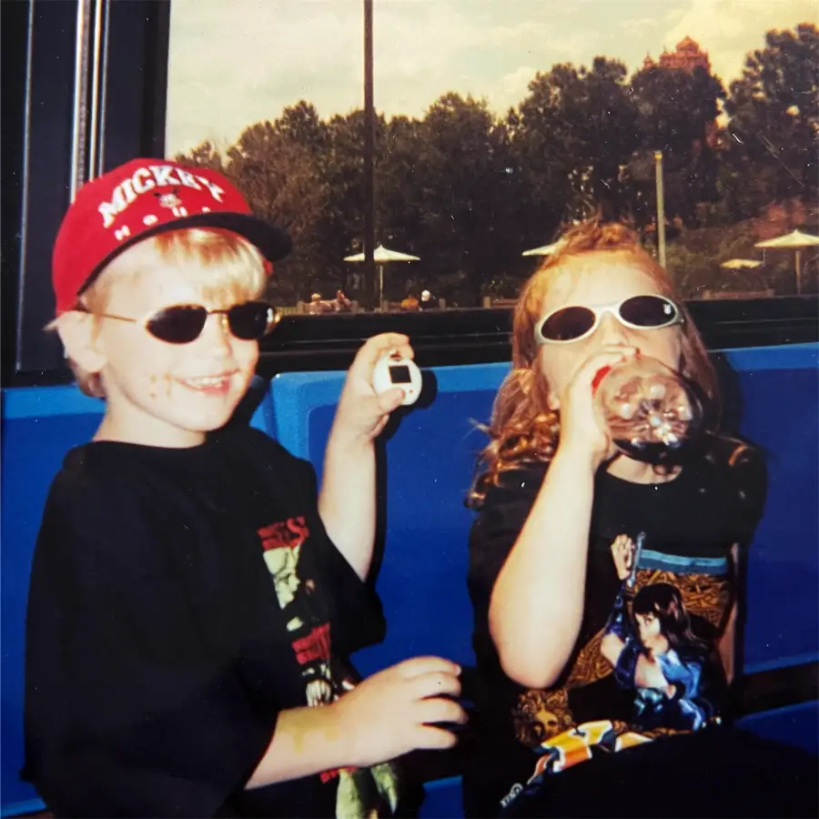 Two young children wearing sunglasses and black t-shirts sit on a blue bench. The boy on the left wears a red cap and smiles while holding a small object. The girl on the right drinks from a soda bottle. Trees and umbrellas are in the background.