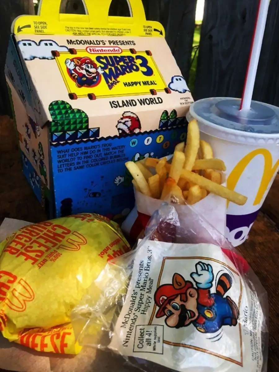 A McDonald's Super Mario Bros. 3 Happy Meal box is shown with fries, a soft drink, a cheeseburger, and a toy in vintage packaging featuring Mario graphics, all set on a wooden surface.