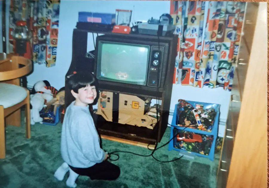 A child kneels on green carpet in front of a vintage TV playing a video game, with colorful sports-themed curtains and toys nearby in a cozy, retro-style room.