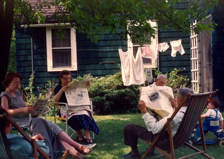 Four adults sit on lawn chairs in a shaded backyard, reading newspapers. Laundry hangs on a line behind them, and a small child sits in a stroller nearby. A dark green house and trees are in the background.