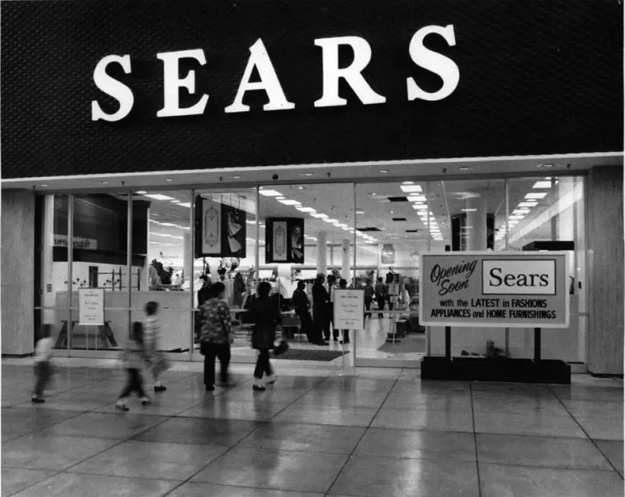 Black and white photo of a Sears store entrance in a mall. People are walking in and out. A sign near the door reads, “Opening Soon: Sears with the latest in fashions, appliances and home furnishings.”