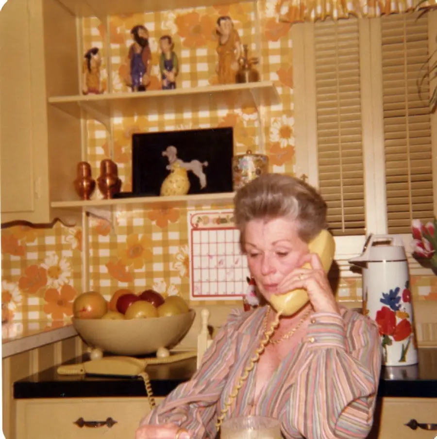 A woman with short gray hair sits in a vintage kitchen with yellow plaid wallpaper, talking on a yellow rotary phone. Behind her are fruit, decorative items, and a wall calendar.