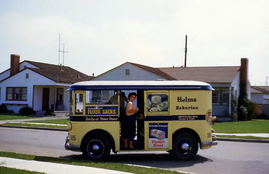 A vintage yellow Helms Bakeries delivery truck is parked on a suburban street, with a woman standing at the open side door. Two single-story houses with lawns are visible in the background.