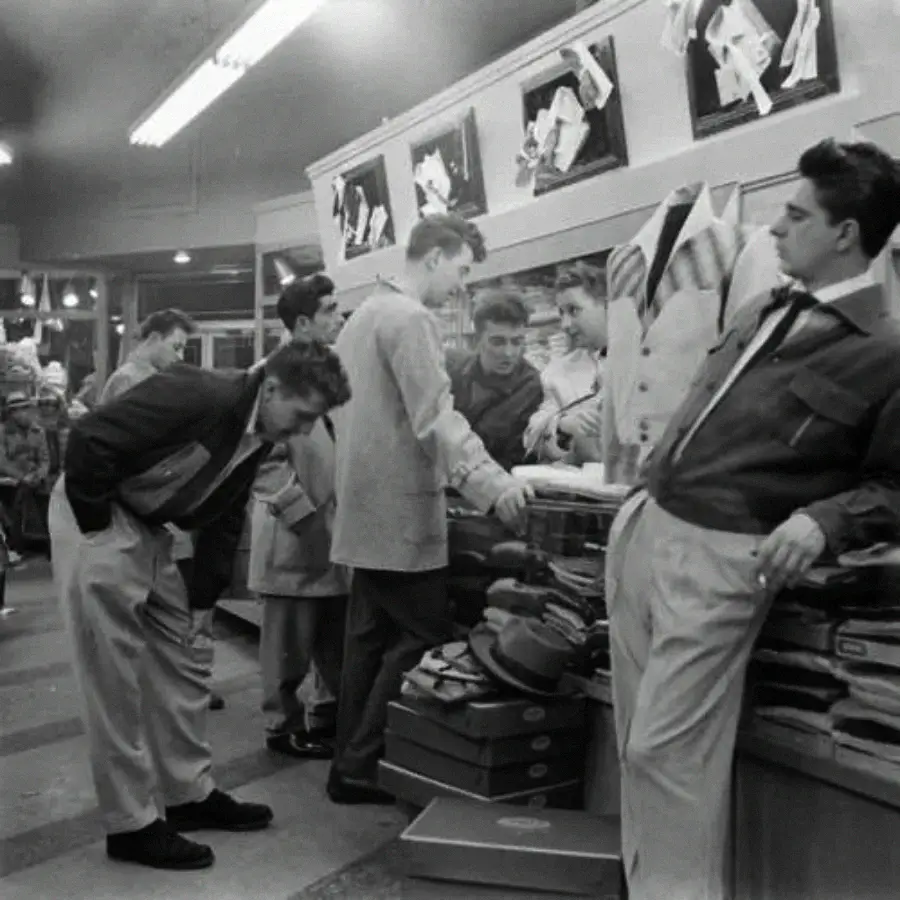 A group of young men in 1950s-style clothing gather in a store, examining hats and jackets displayed on shelves, with framed pictures on the wall above them.