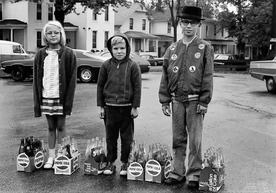 Three children stand on a street with vintage houses and cars behind them. Each child stands in front of glass soda bottles in cardboard carriers labeled Pepsi and Coca-Cola. The boy in the center looks grumpy.