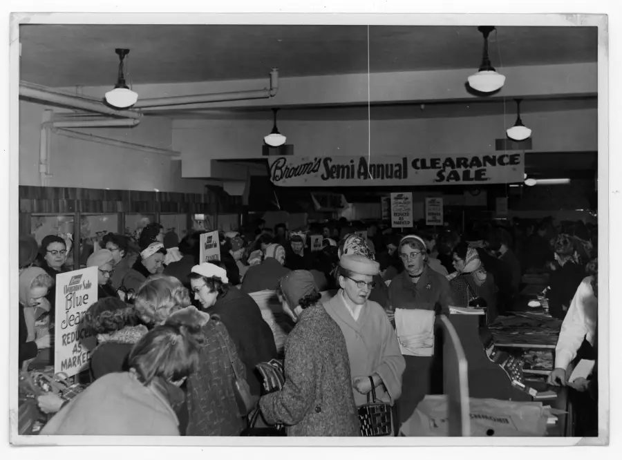 A crowded store filled with shoppers during a semi-annual clearance sale; many people wearing coats and hats, with sale signs and merchandise visible throughout the busy scene.