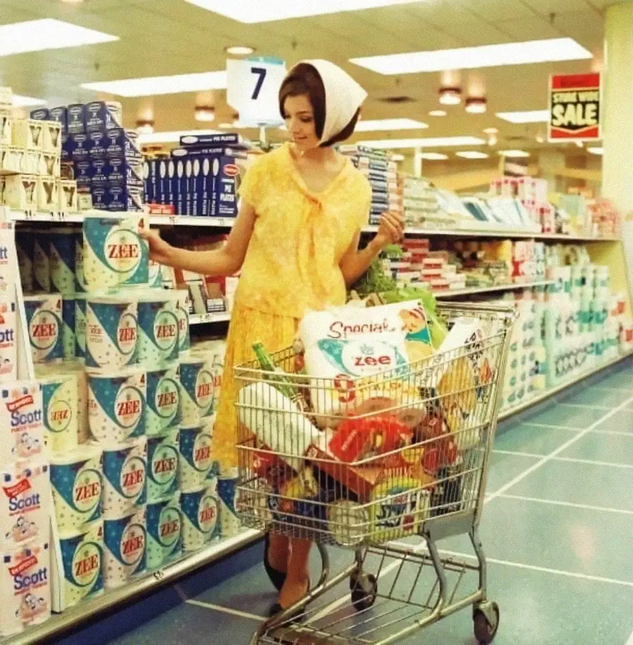 A woman in a yellow dress shops in a vintage supermarket, browsing paper goods. Her cart is full of groceries. The store shelves display products like Scott and ZEE napkins. The scene appears to be from the 1960s.