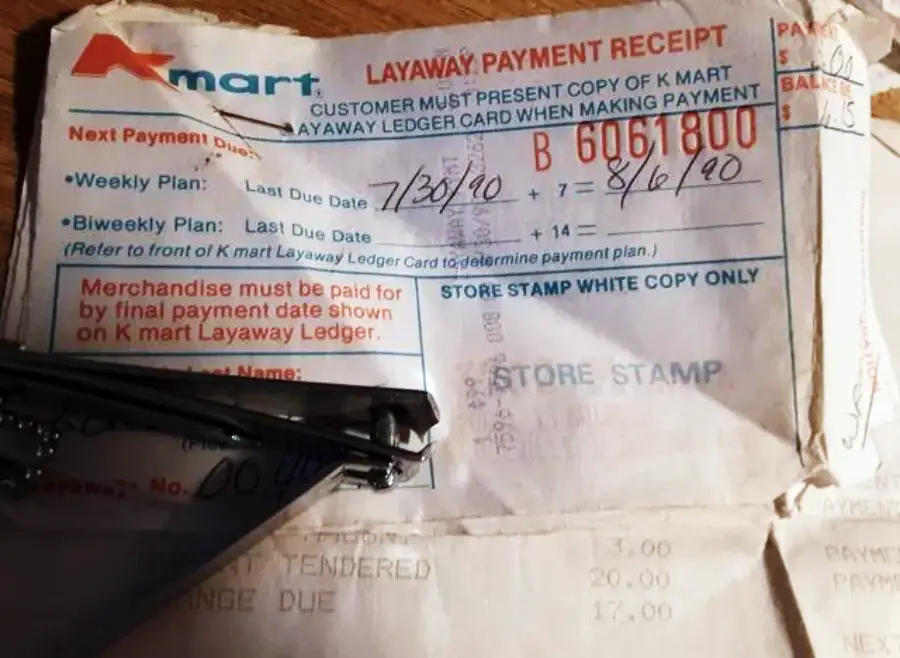 A vintage Kmart layaway payment receipt from 1990 shows handwritten payment dates and amounts, with some of the text faded. Part of a black object and another paper partially cover the receipt.
