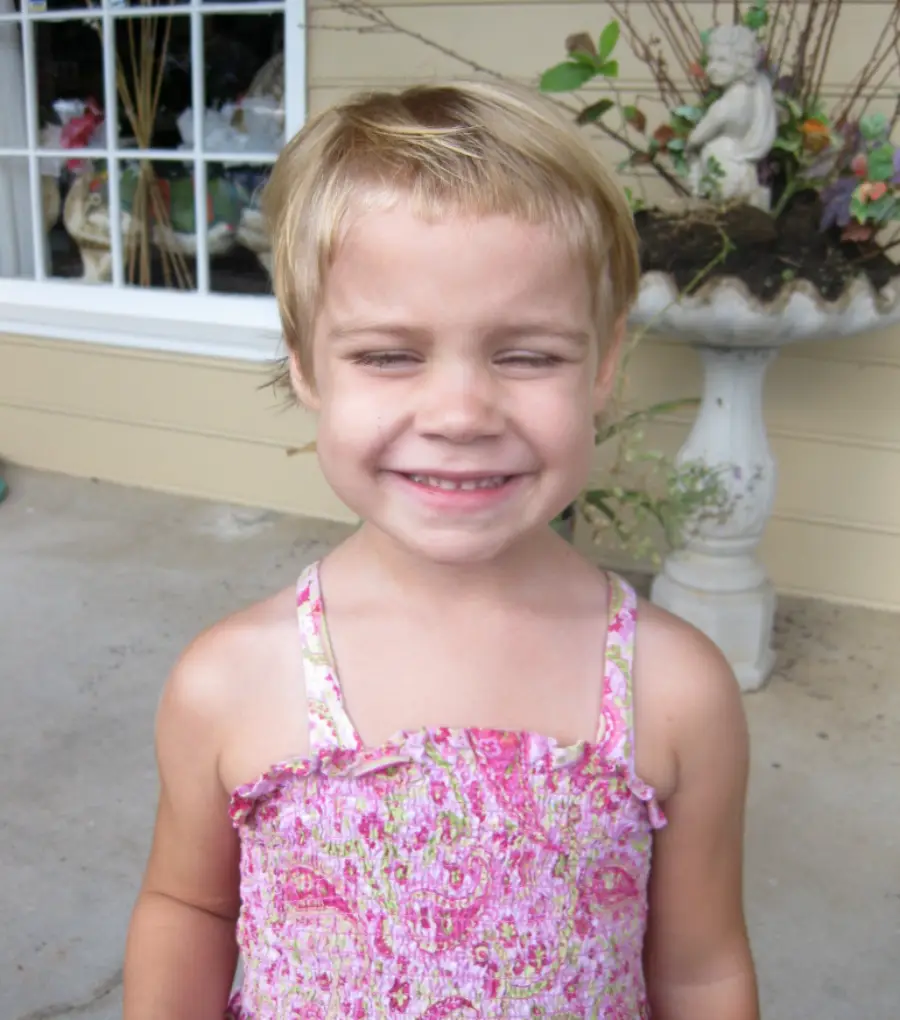 A young child with short blonde hair smiles with eyes closed, wearing a pink patterned dress, standing outside near a large planter and a window with decorative plants visible in the background.