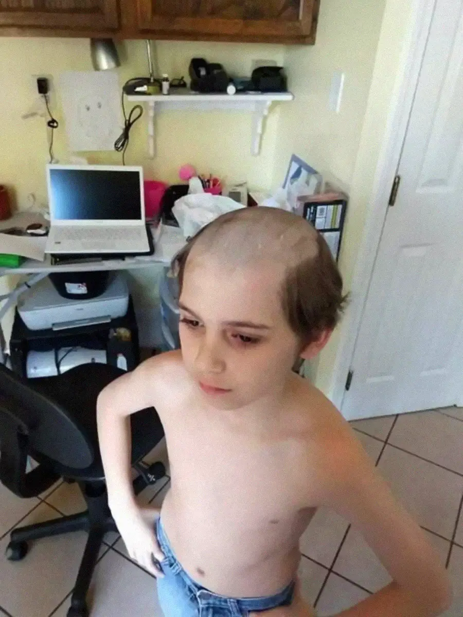 A shirtless child with their hands on their hips stands in a room with a computer desk behind them; their hair has been shaved off on top, leaving hair only around the sides and back of their head.
