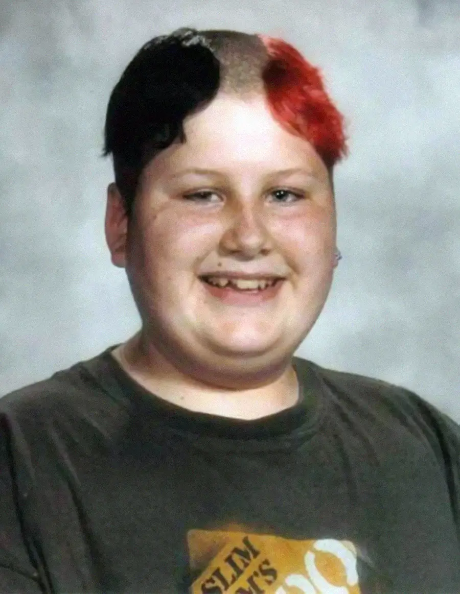 A smiling person with very short hair, dyed black on one side and red on the other at the front, wears a dark t-shirt with a partially visible graphic against a gray studio background.