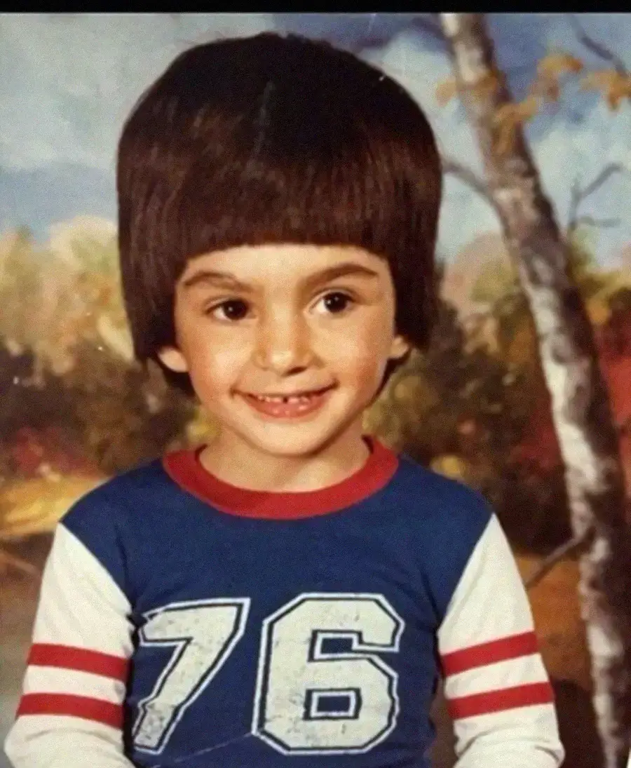 A young child with a large bowl haircut smiles at the camera, wearing a blue shirt with white and red stripes on the sleeves and the number 76 on the front. The background shows an autumnal outdoor scene.