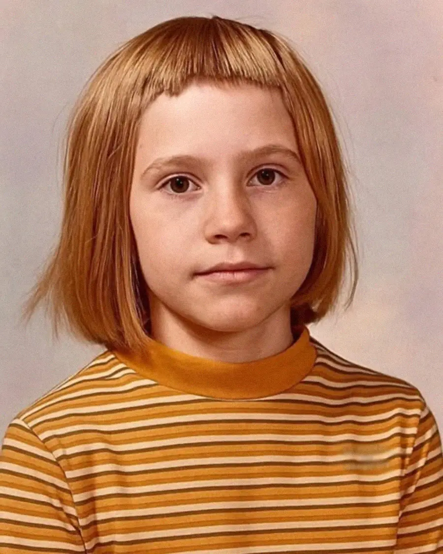 A young child with straight light brown hair and short bangs, wearing a yellow and white striped shirt, poses for a school photo against a neutral background.