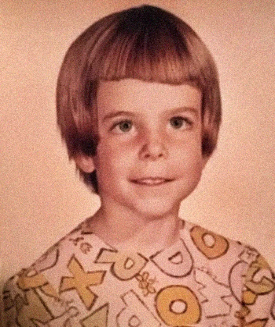 A young child with light brown hair in a bowl cut smiles slightly at the camera. The child is wearing a light-colored shirt with colorful, large letters printed on it. The background is a neutral solid color.