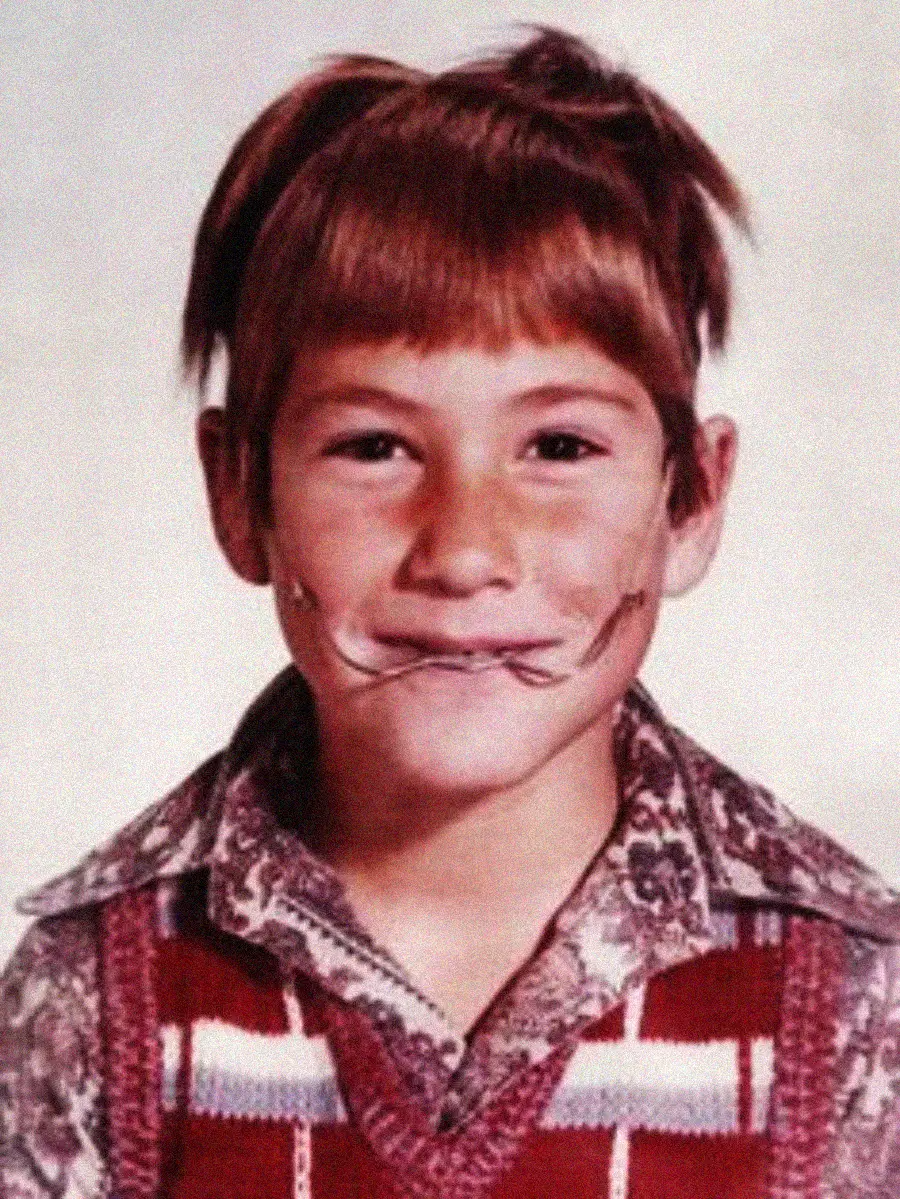 A young boy with brown hair and bangs smiles at the camera. He is wearing a patterned shirt and a red vest. There is a drawn-on mustache and smiley face on his cheeks, resembling doodles with a pen.