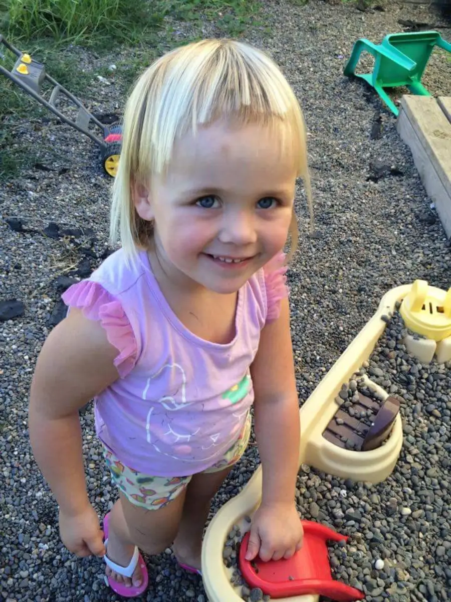 A young girl with blonde hair and short bangs, wearing a light purple shirt and patterned shorts, stands on gravel outdoors, smiling beside a beige water play table with toys.