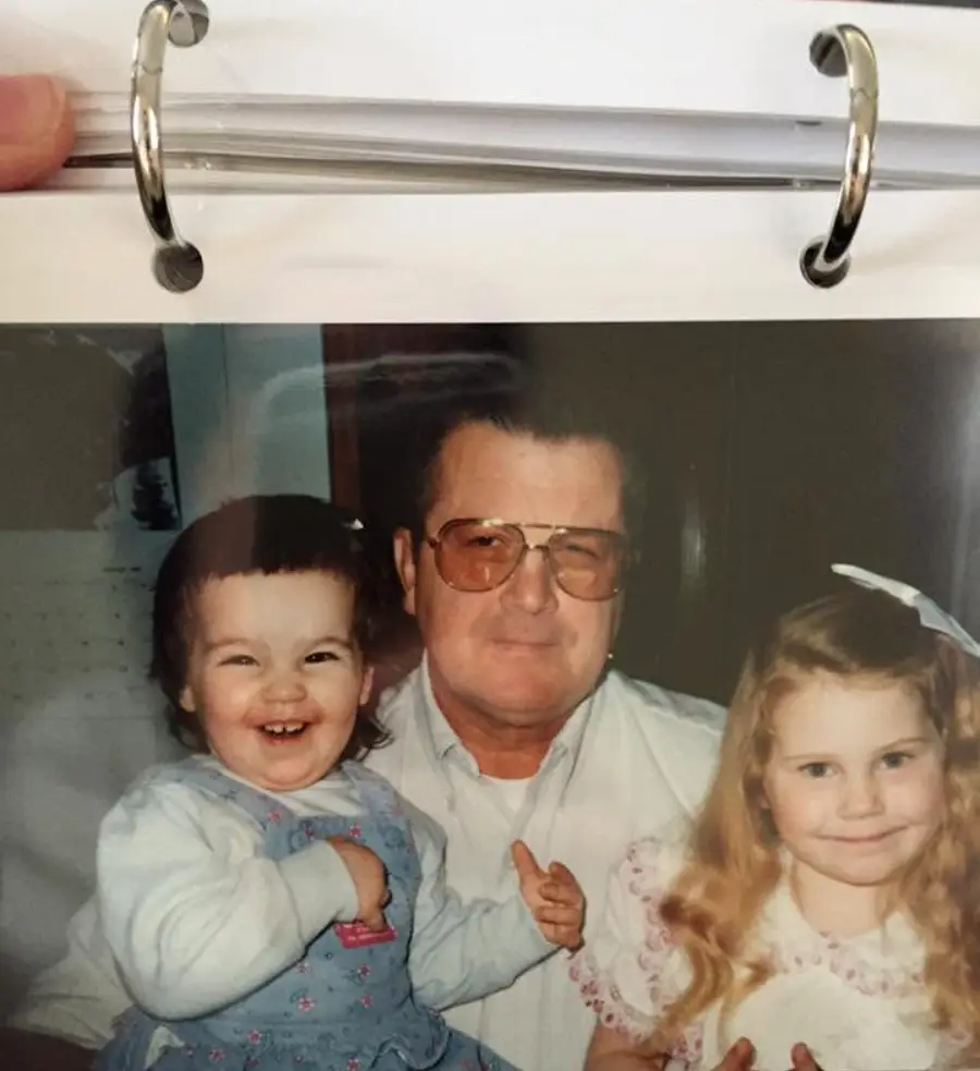 A man wearing glasses sits between two young smiling girls, one with short dark hair and the other with long blond hair and a white bow. They are posing together for a photo, held in a binder.