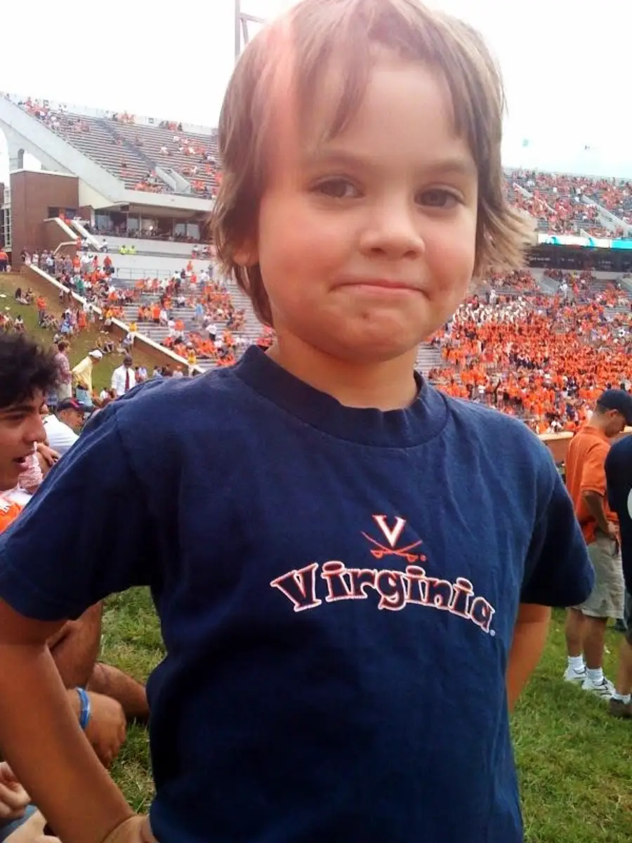 A young child with brown hair stands on grass at a stadium, wearing a navy blue “Virginia” T-shirt. The stands behind are filled with fans, many in orange, suggesting a sporting event.