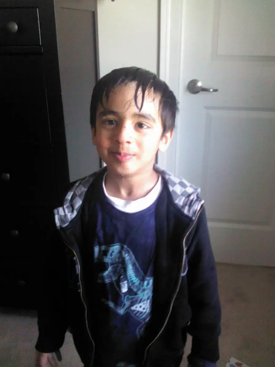 A young boy with wet hair stands indoors, smiling softly. He is wearing a dark hoodie over a graphic t-shirt and is in front of a white door and a black dresser. Light comes in from the side.