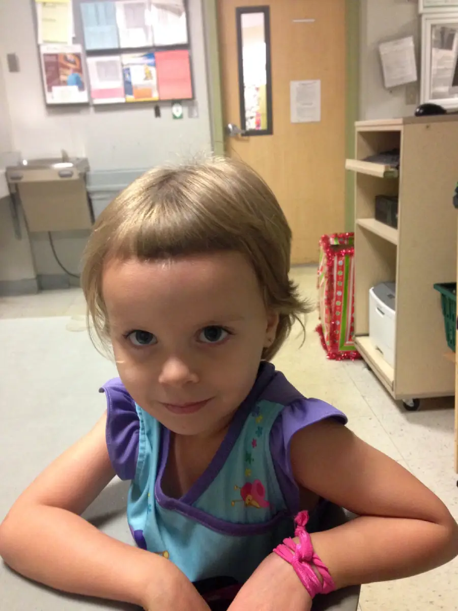 A young child with short, light brown hair and bangs sits at a table, looking up and smiling slightly. The child is wearing a colorful shirt and a pink string bracelet, with an indoor office or classroom in the background.