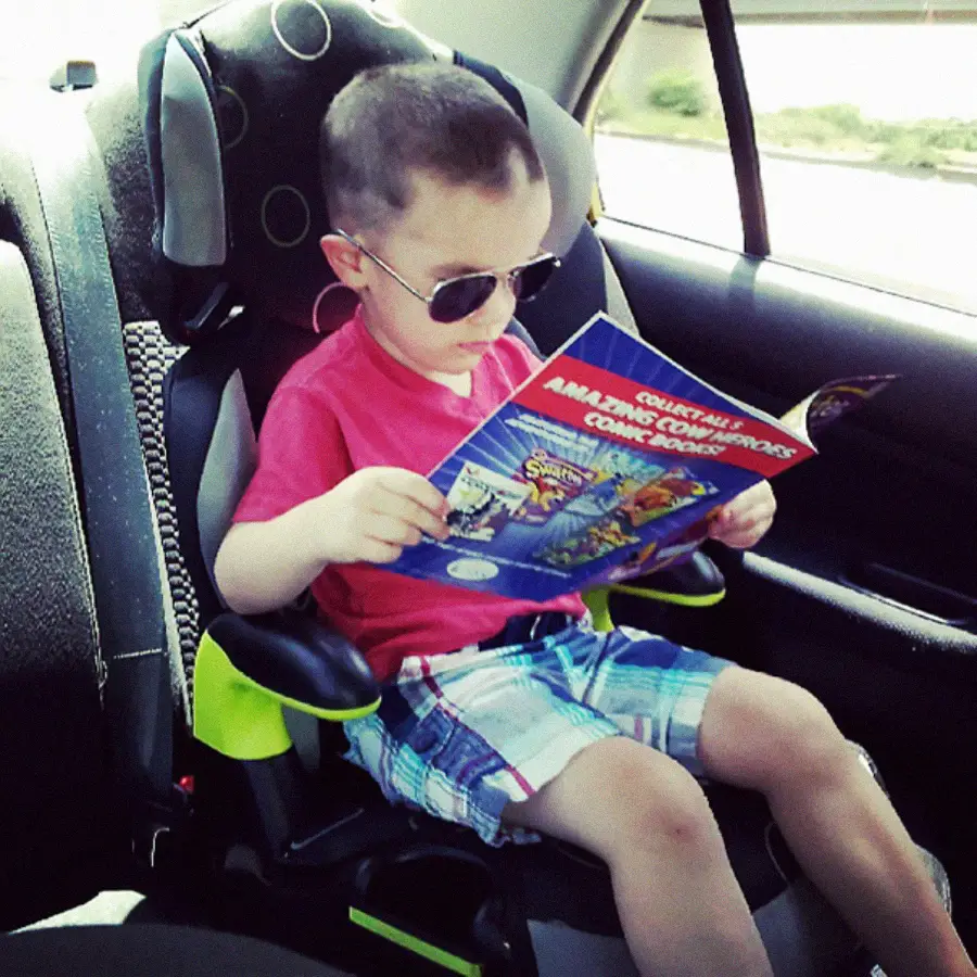 A young boy wearing sunglasses sits in a car seat in the back of a car, reading a colorful comic book. He is dressed in a red shirt and plaid shorts, and sunlight streams through the window.