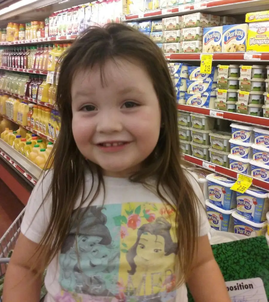 A young girl with long brown hair smiles while standing in a grocery store aisle. She is wearing a white shirt with cartoon princesses on it, and cartons of juice and tubs of margarine are visible on the shelves behind her.