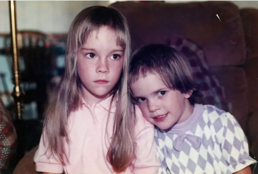 Two young girls sit close together on a couch. The girl on the left has long, straight hair and a serious expression, while the younger girl on the right with short hair leans in smiling, wearing a patterned top with a bow.