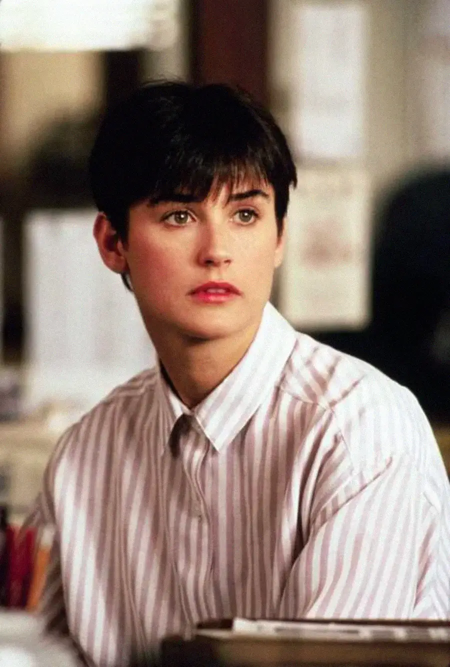 A person with short dark hair and a light complexion wears a light-colored, striped button-up shirt. They are indoors, seated, and looking slightly to the side with a neutral expression.