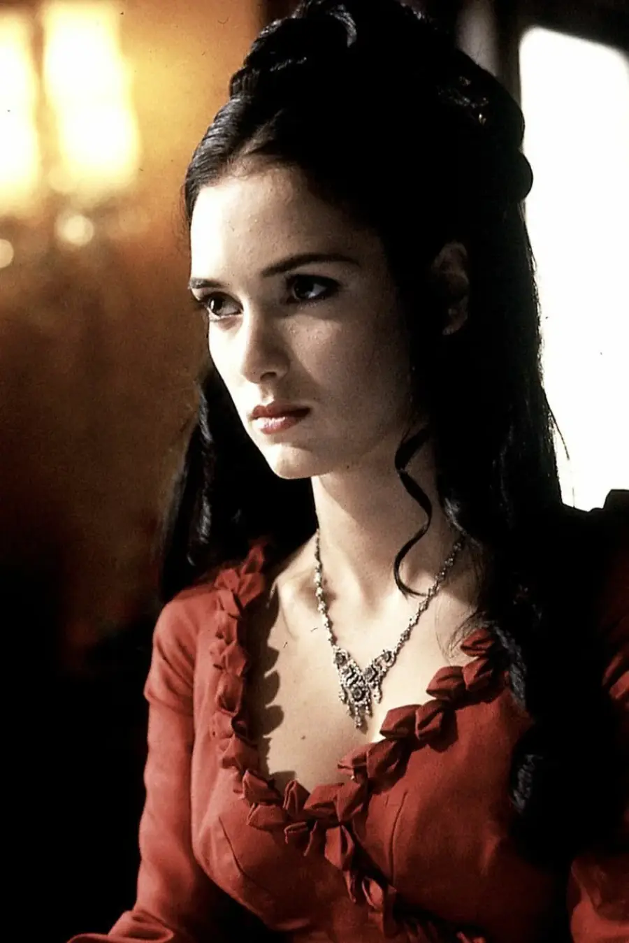 A woman with long dark hair wears a red dress with ruffle details and a decorative necklace, sitting indoors with a serious expression. The background is softly lit.