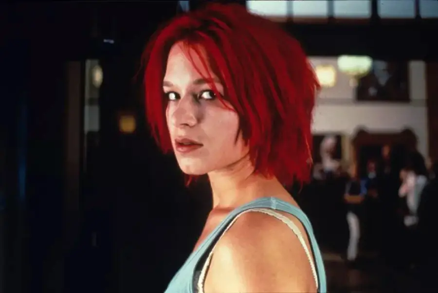 A woman with bright red, tousled hair and a blue tank top looks over her shoulder toward the camera, standing indoors with a blurred background.