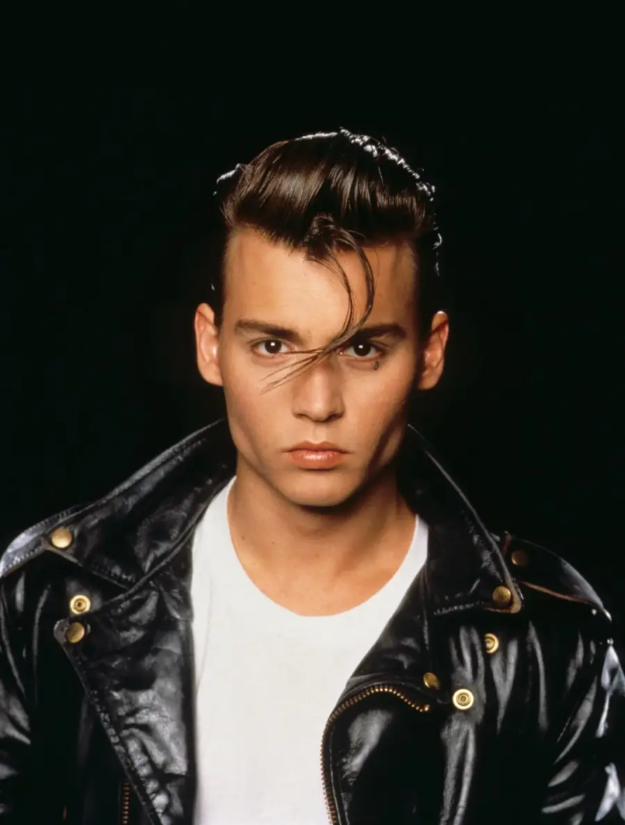 A young person with slicked-back hair and a single strand falling forward wears a black leather jacket over a white T-shirt, looking seriously at the camera against a plain dark background.