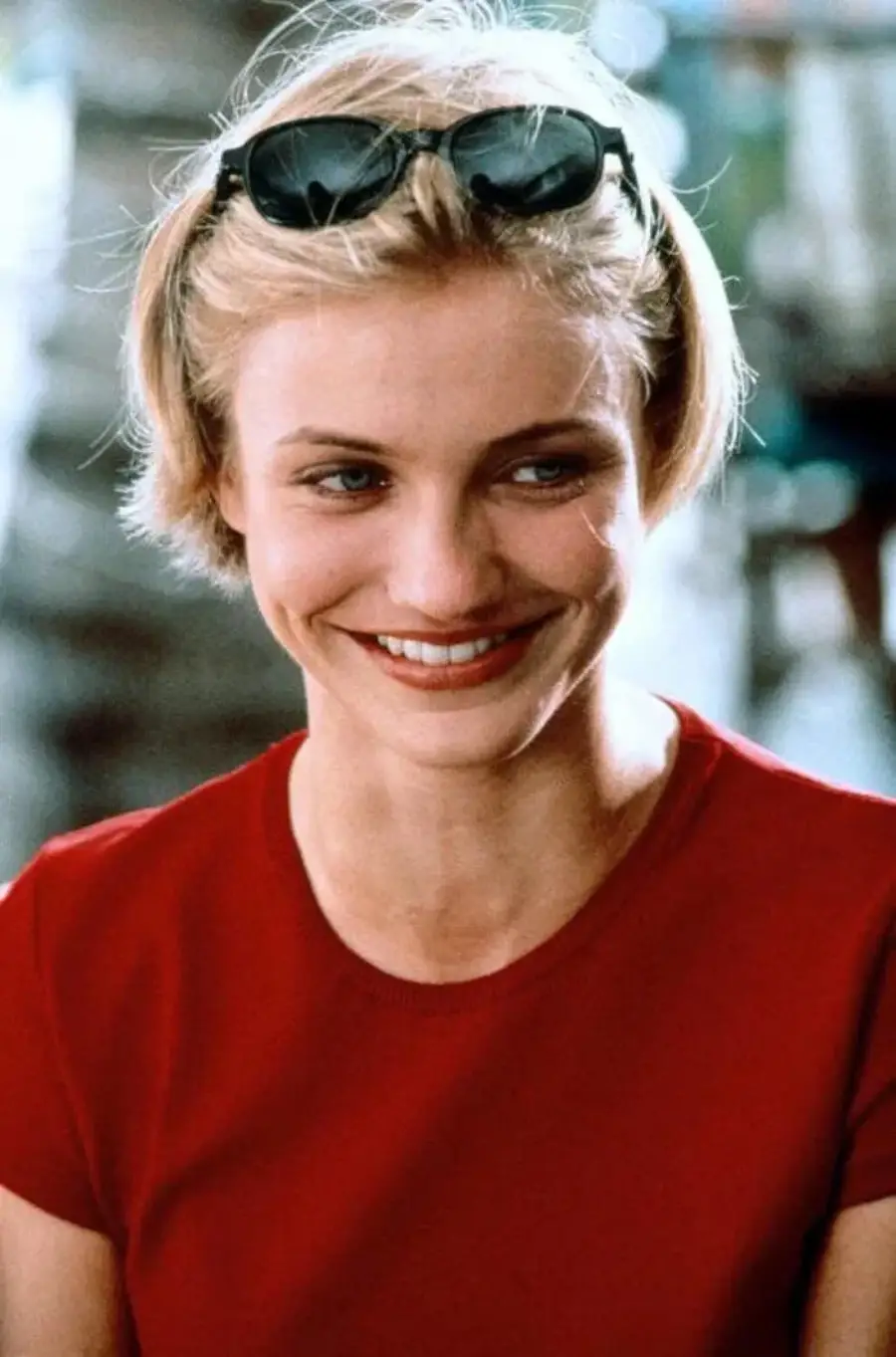 A woman with short blonde hair, wearing sunglasses on her head and a red top, is smiling softly while looking to the side.