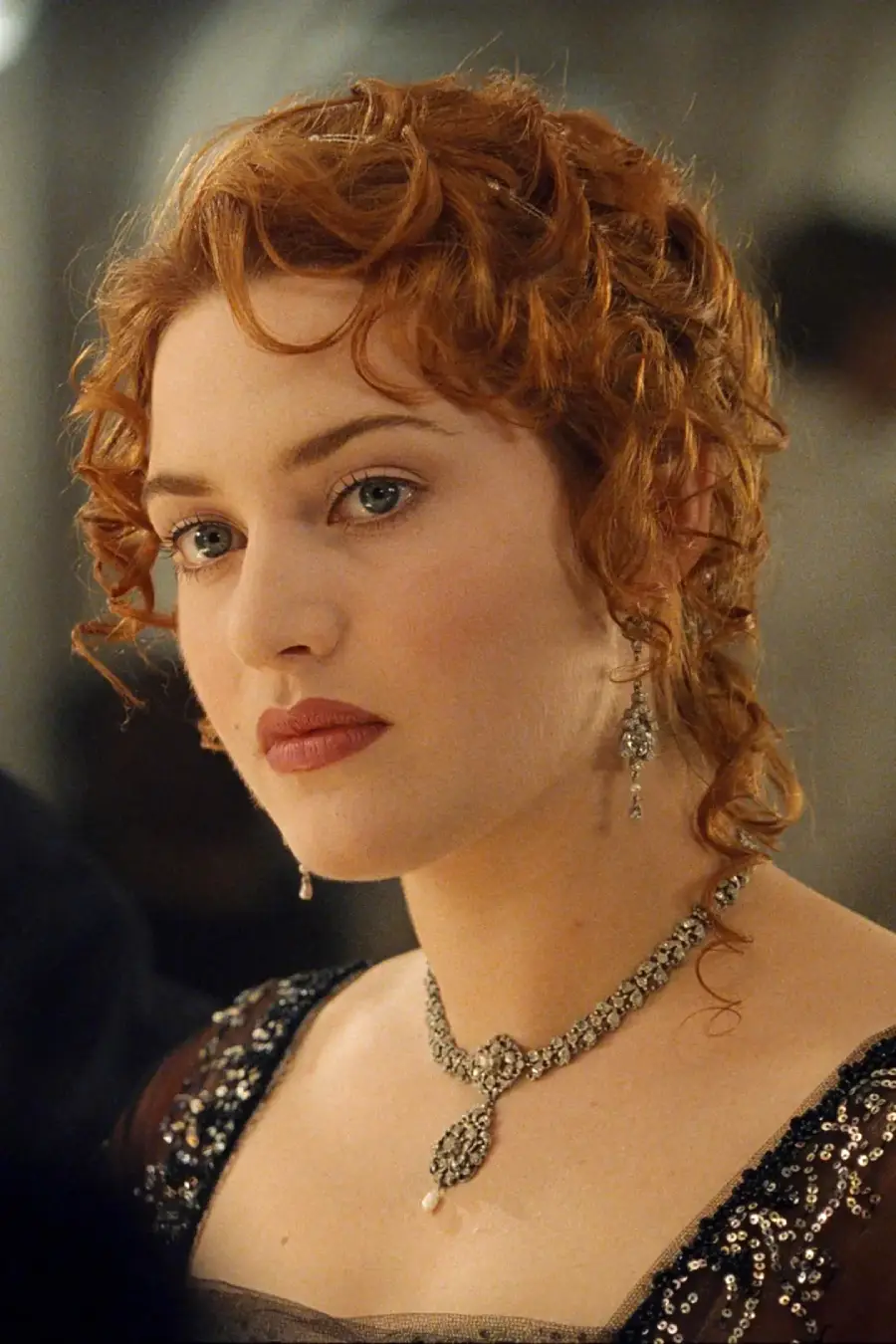 A woman with curly red hair and fair skin wears an ornate necklace, dangling earrings, and a dark, elegant dress. She looks thoughtful and is set against a softly blurred background.