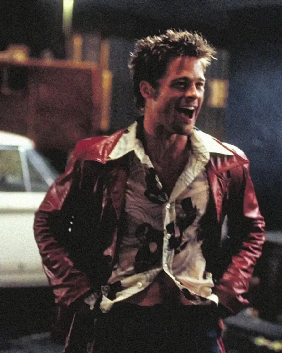 A man with spiky hair wearing a red leather jacket and patterned shirt stands laughing with his hands on his hips in a dimly lit urban setting, with a white car and blurred background behind him.
